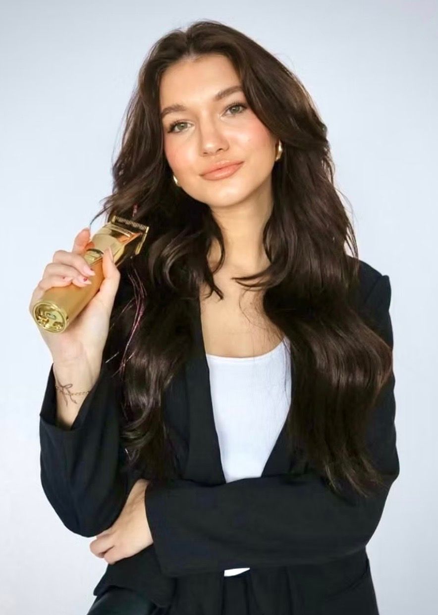 Woman in black blazer holds gold hair clippers, looking at camera.