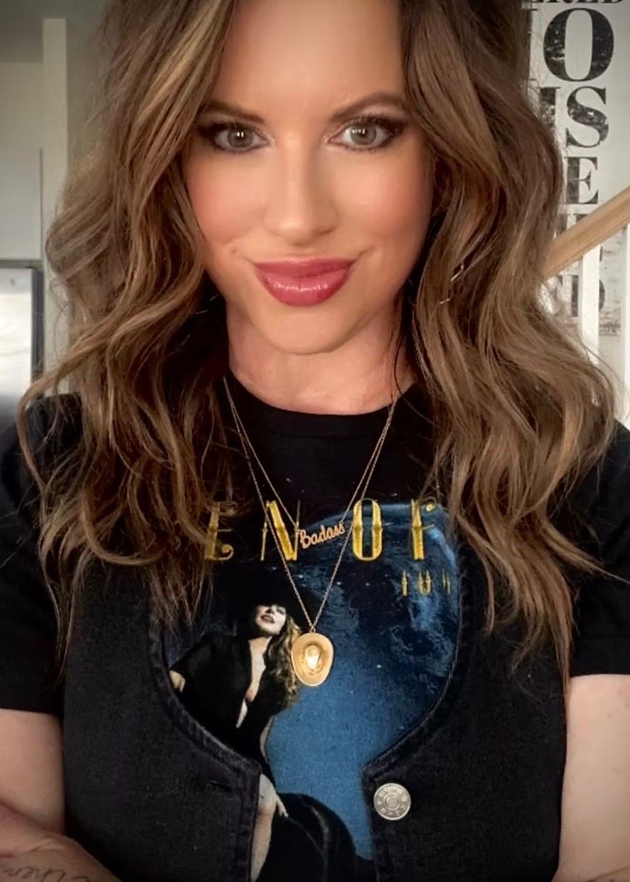Woman with wavy brown hair, wearing a black vest over a band t-shirt, smiling, in front of a staircase.