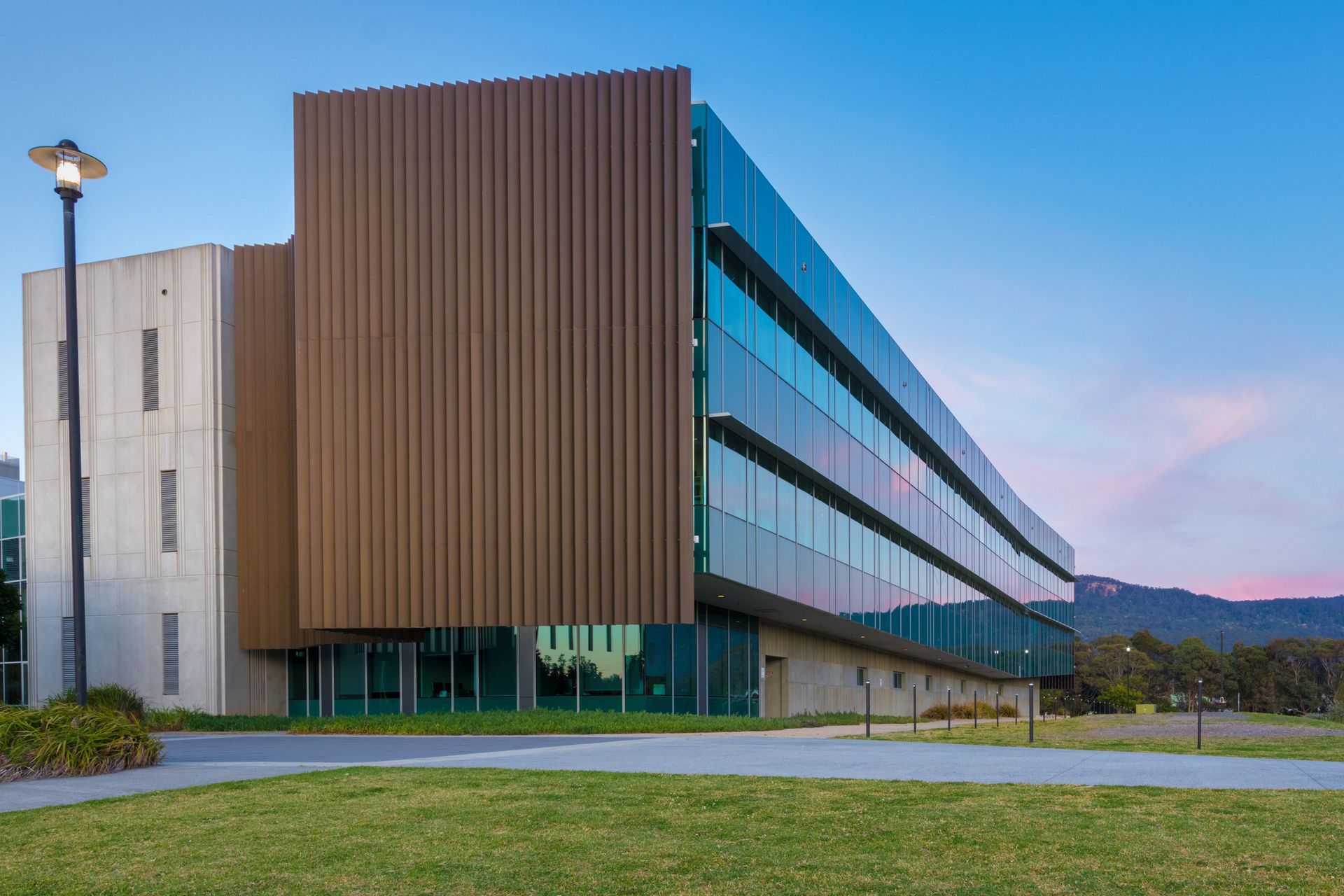 Modern university building with brown, vertical panels, glass facade, and grassy lawn.