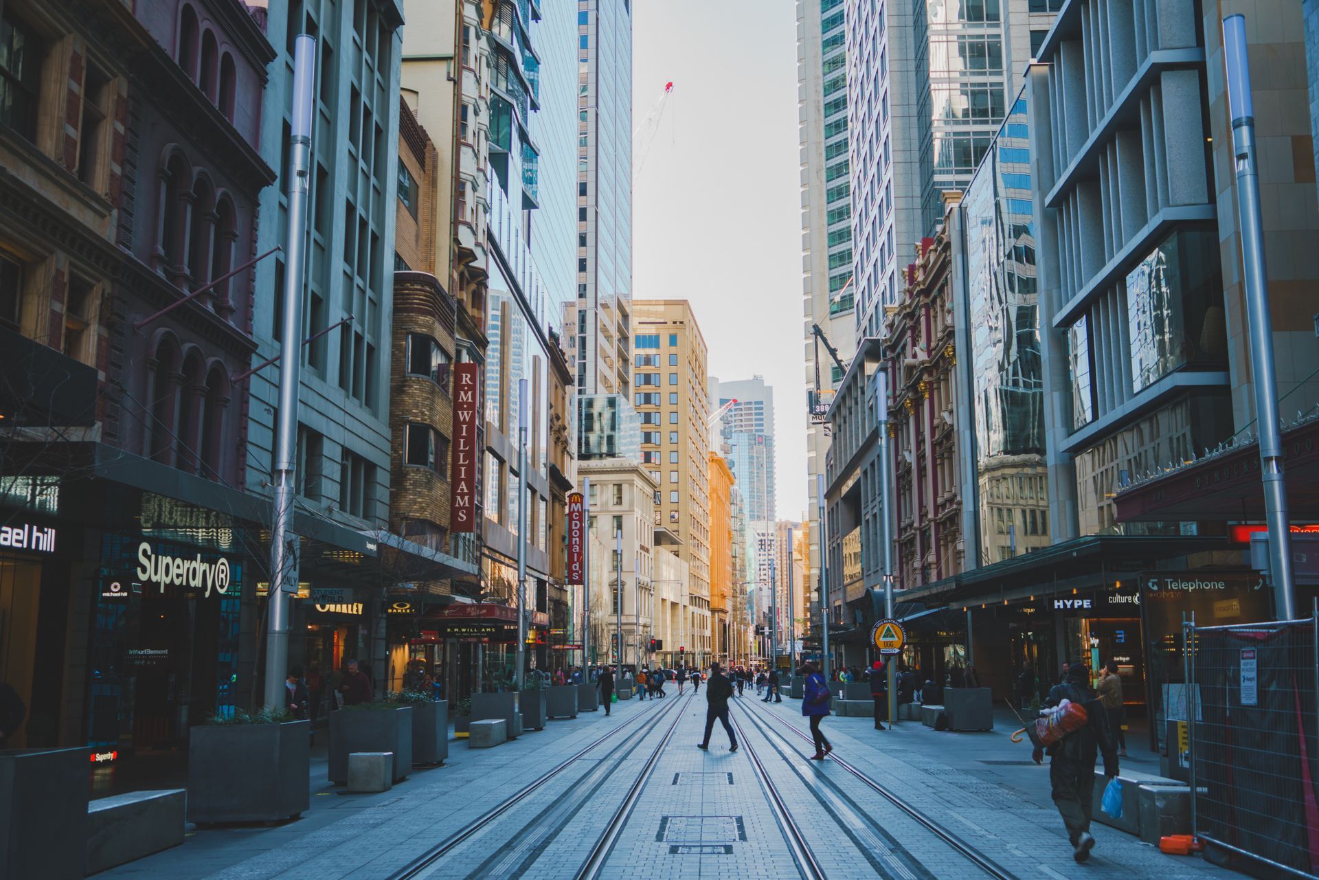 City street lined with tall buildings. People walk on the street and cross the train tracks.