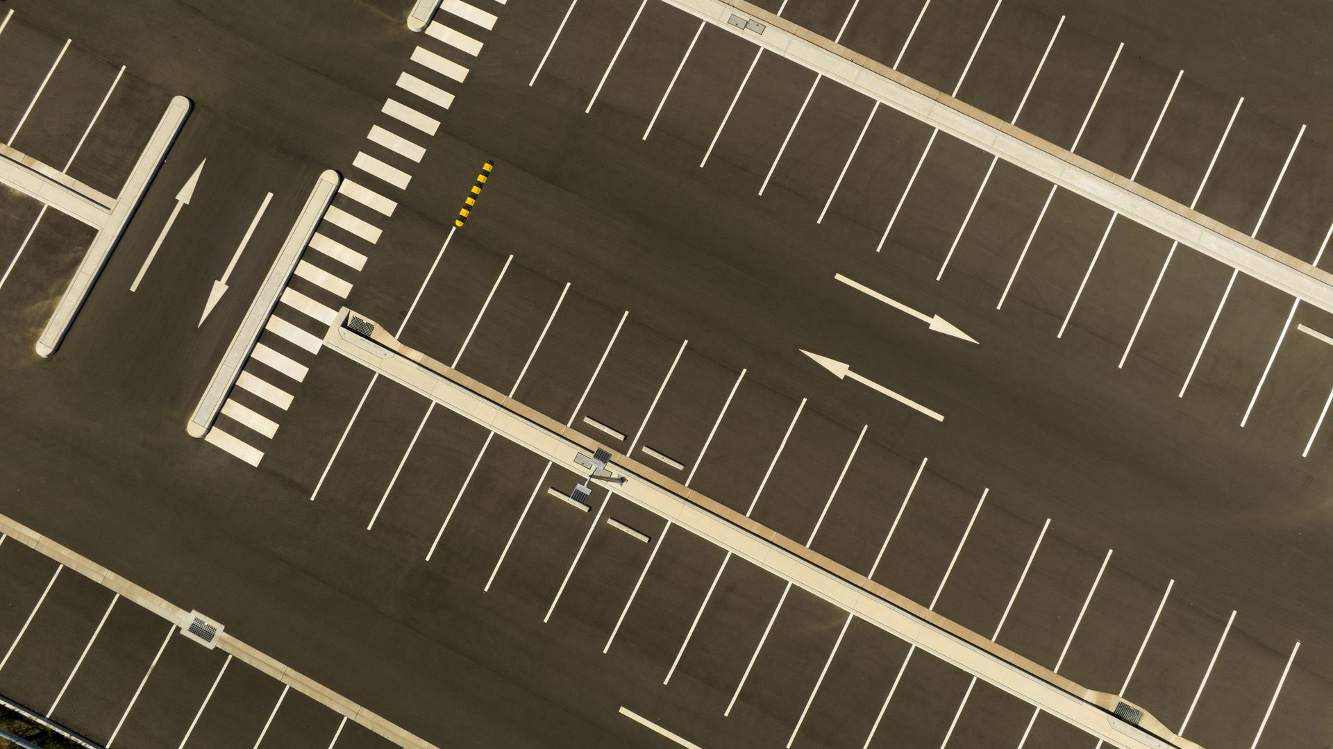 Overhead view of an empty parking lot with marked spaces and crosswalks. Asphalt is dark, markings are white.