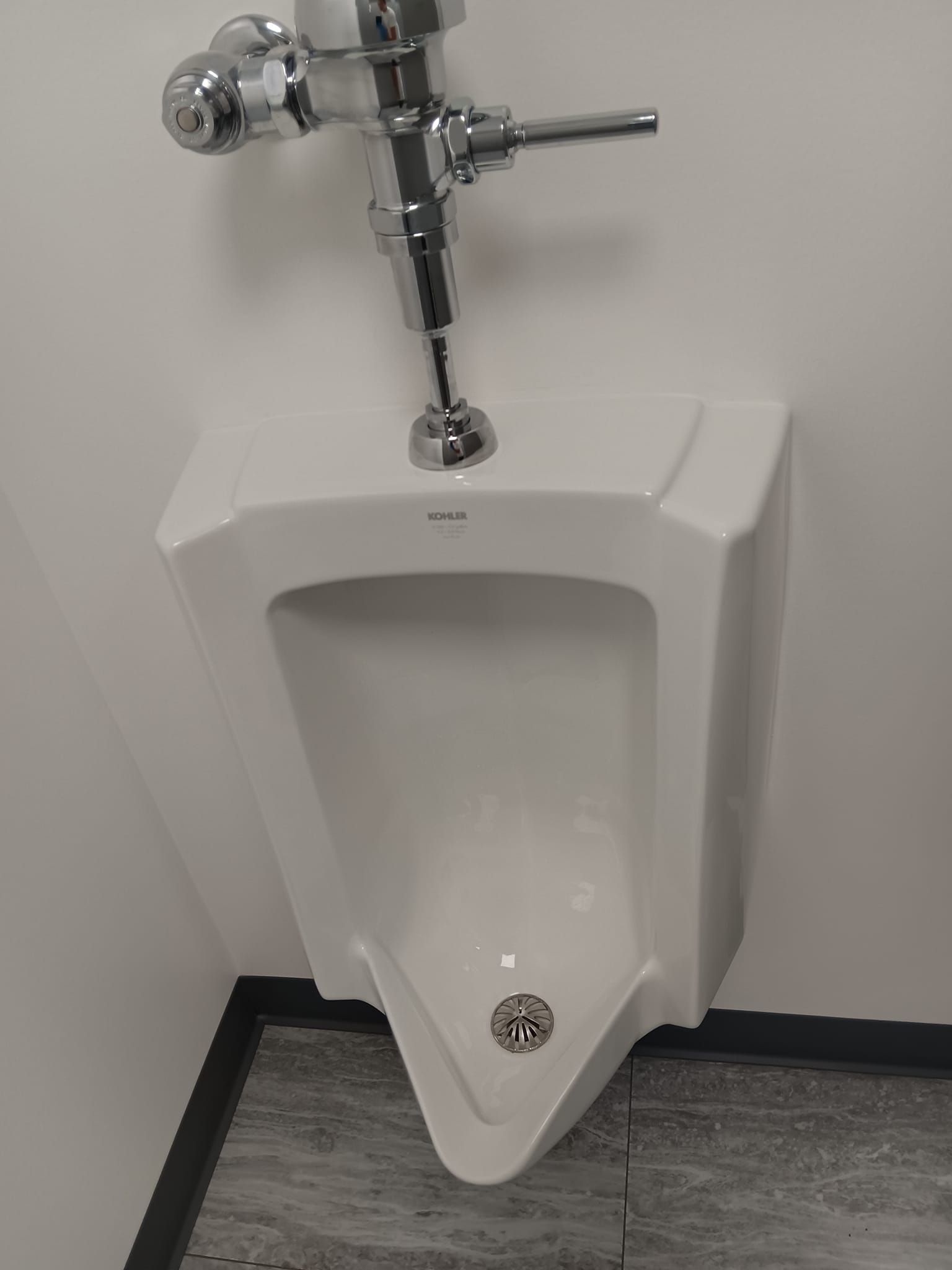 A white urinal is sitting in a bathroom next to a white wall.