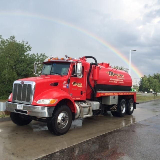 A red vacuum truck is parked on the side of the road with a rainbow in the background.