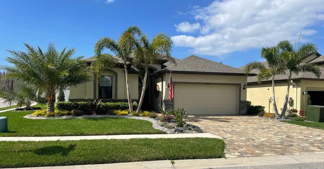 Tan house with brown roof, palm trees, and green grass. Brick driveway on a sunny day.
