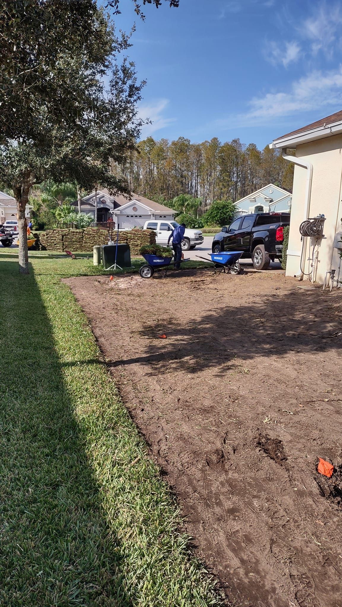 Workers preparing a dirt bed for landscaping near a house and a green hedge.