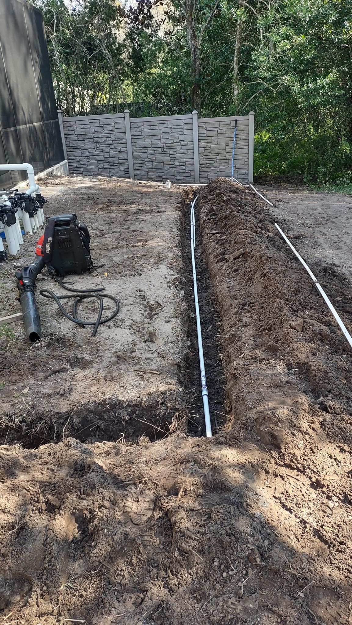 Trench with exposed pipe, equipment nearby. Dirt path, fence, and trees in the background.