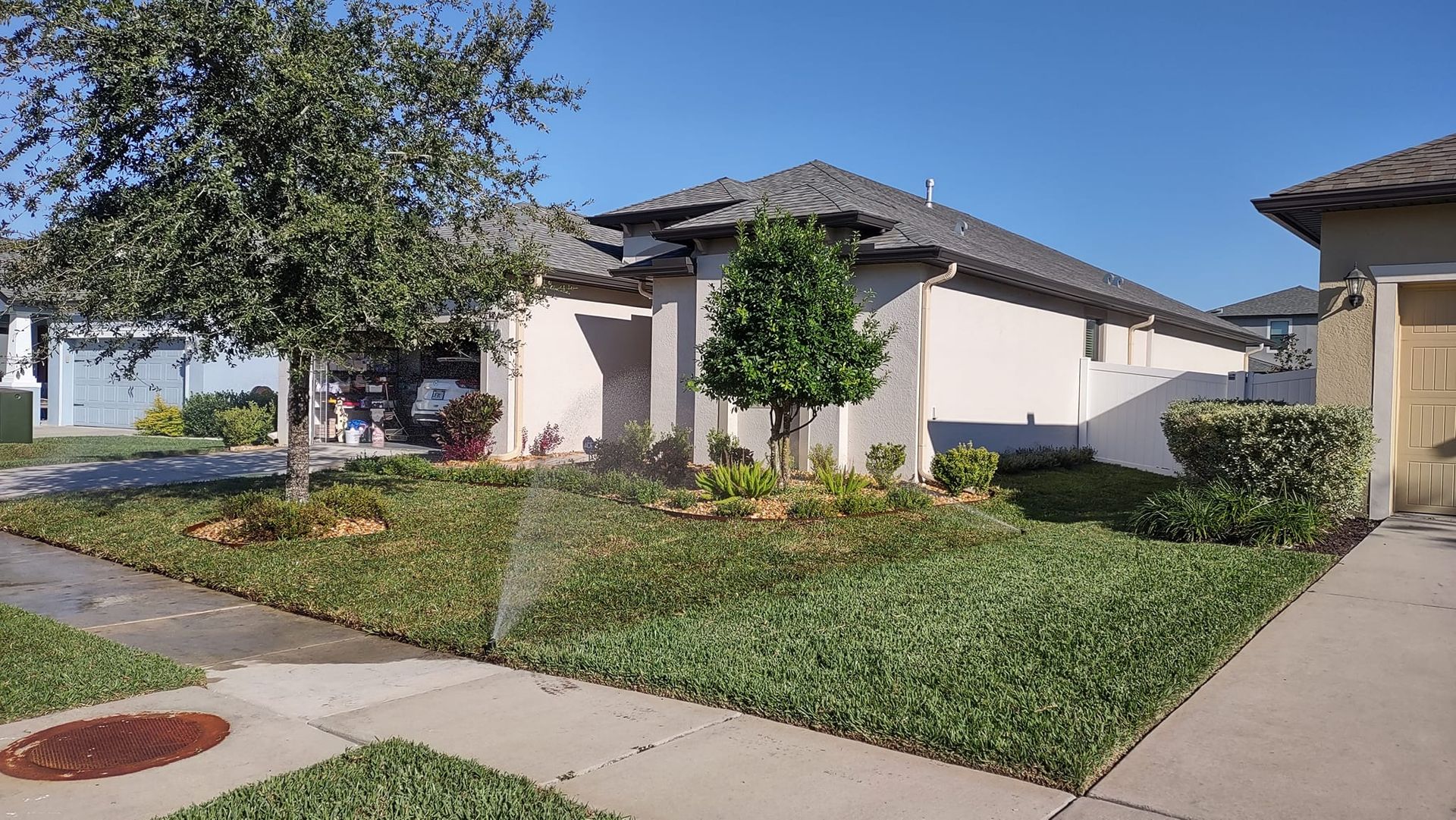 Suburban house with green lawn, sprinklers, and trees; sunny day.