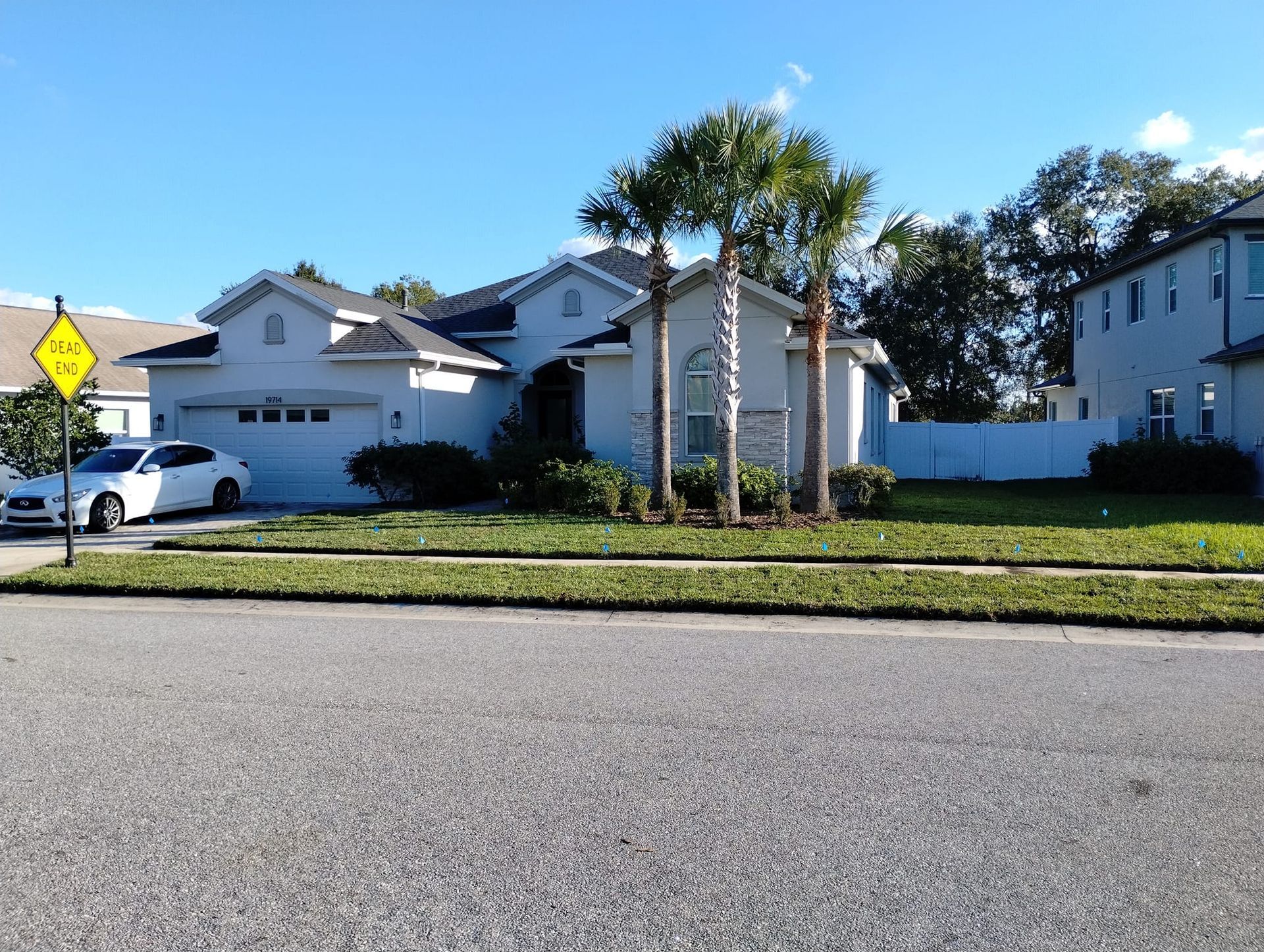 A single-story, light-colored house with a two-car garage, palm trees, and green lawn. A white car is parked in the driveway.