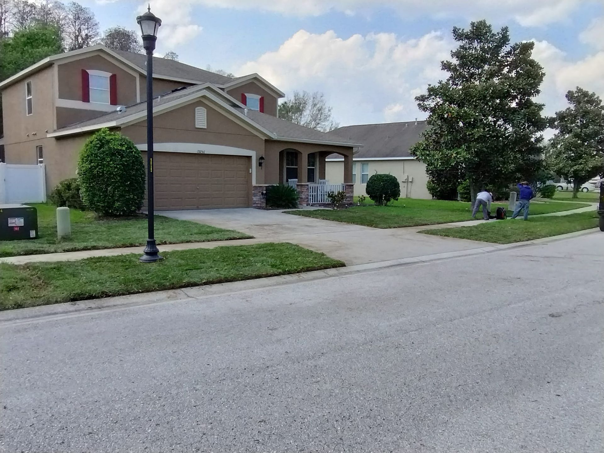 Suburban house with brown facade, green lawn, and people working in the yard on a sunny day.