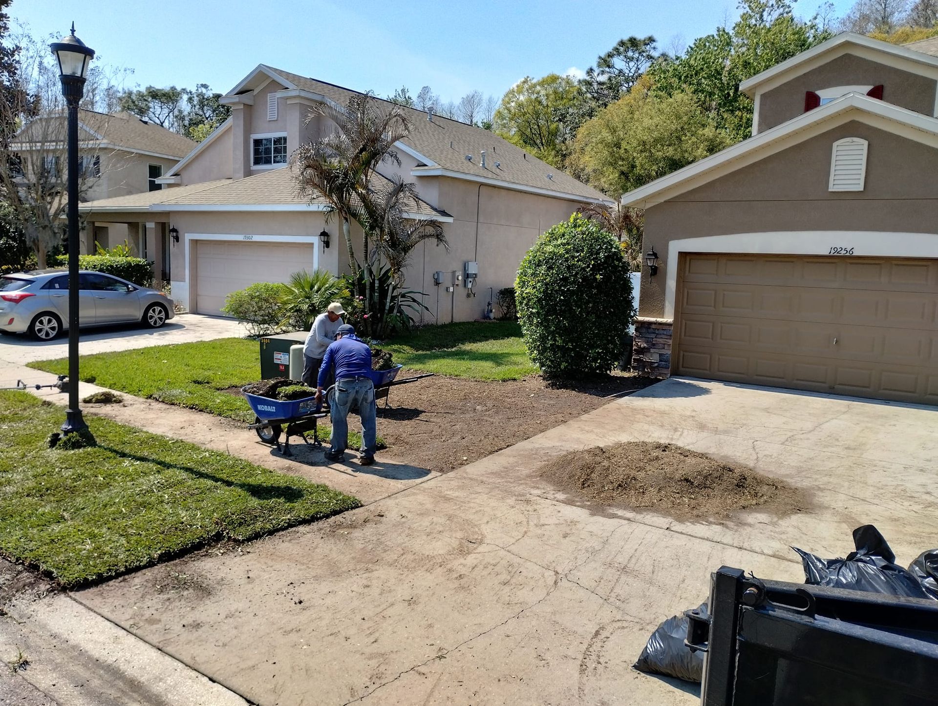 Two people landscaping a front yard, brown soil, blue cart. Houses, car, and blue sky in the background.