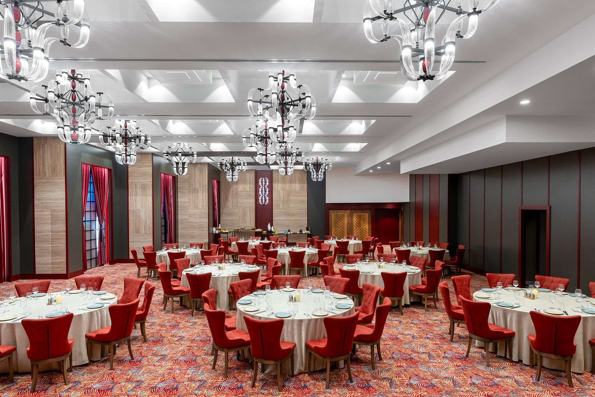 Empty banquet hall with red chairs, round tables, patterned carpet, and chandeliers ceiling lights
