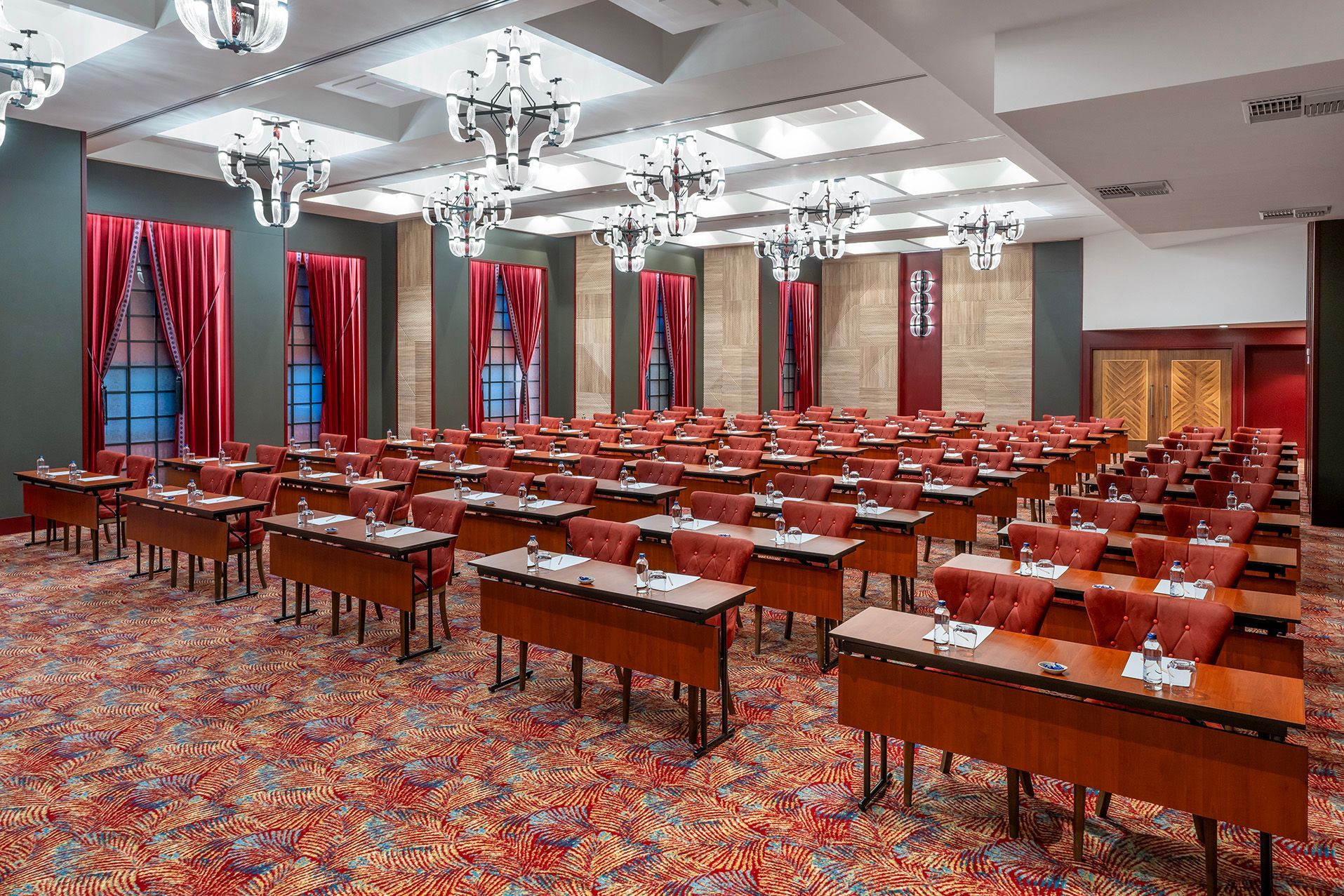 Empty conference room with rows of tables and red chairs under chandeliers, carpeted floor, and tall windows