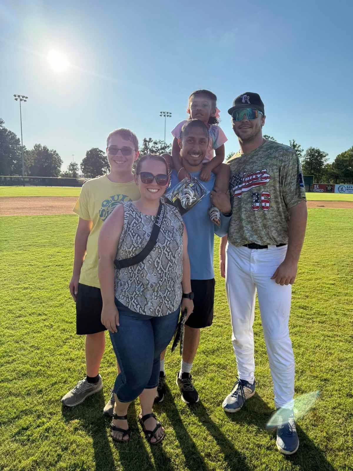 Five people pose for photo on a baseball field. Sunny outdoors; green grass.