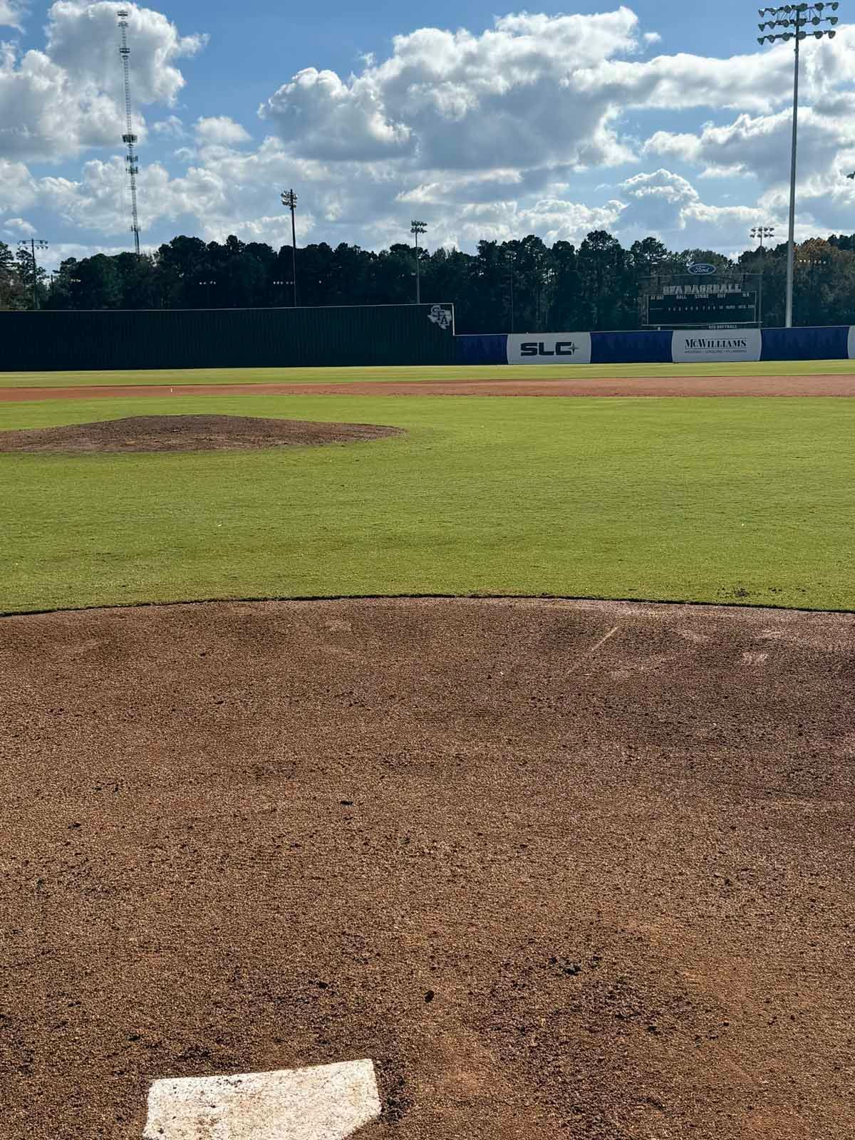 Baseball field with a brown dirt infield and green outfield under a cloudy sky.