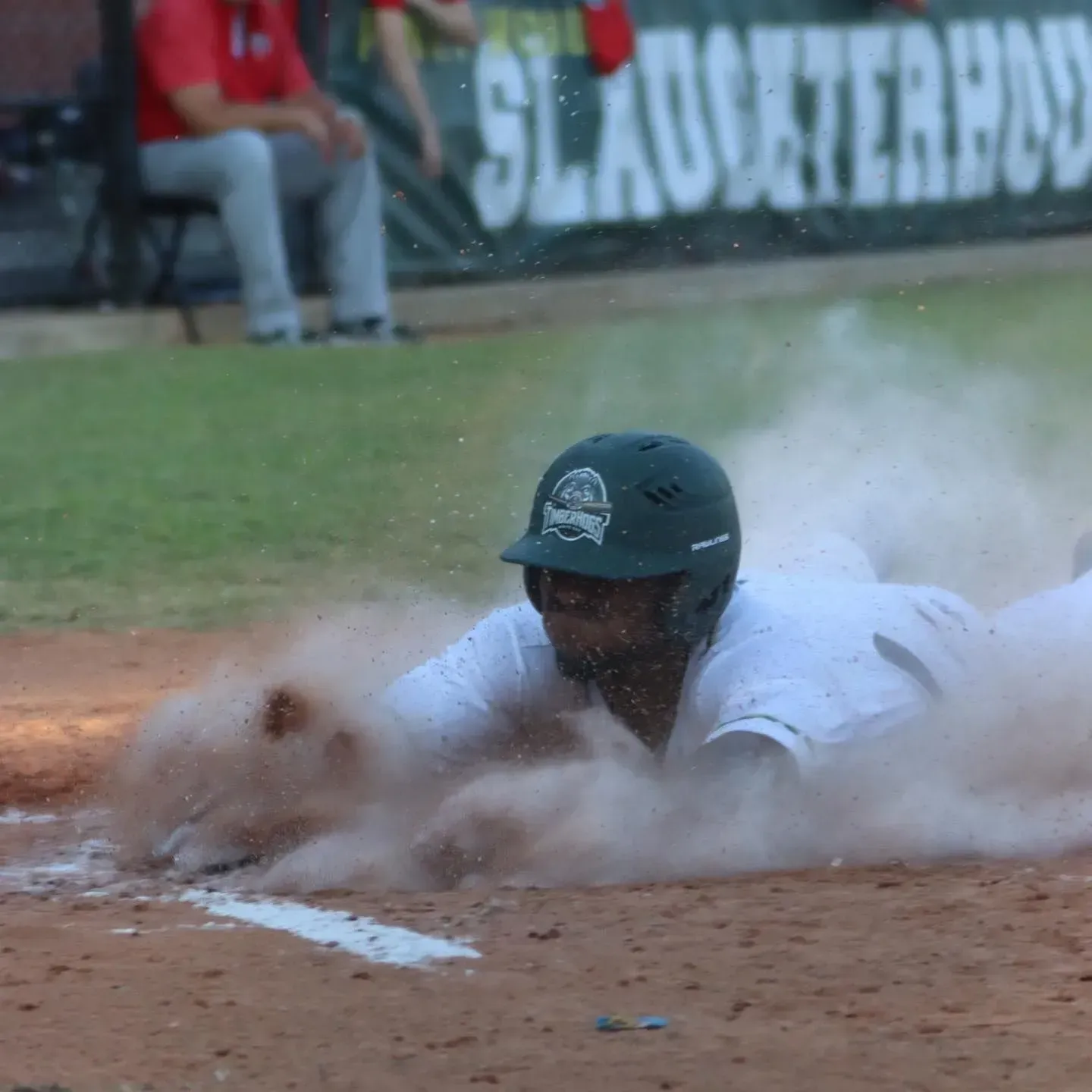 Baseball player sliding into home plate, kicking up dirt. Green helmet, white uniform, baseball field.