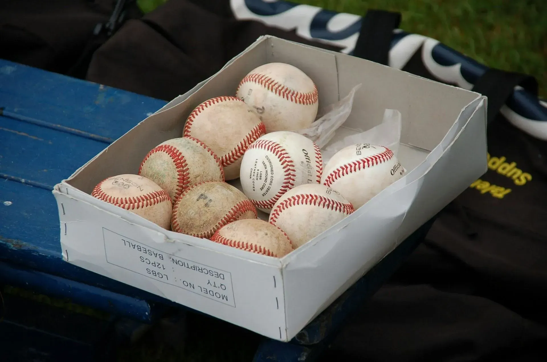 Box of used baseballs on a blue bench, next to a black bag.