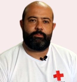 Um homem com barba está vestindo uma camisa branca com uma cruz vermelha