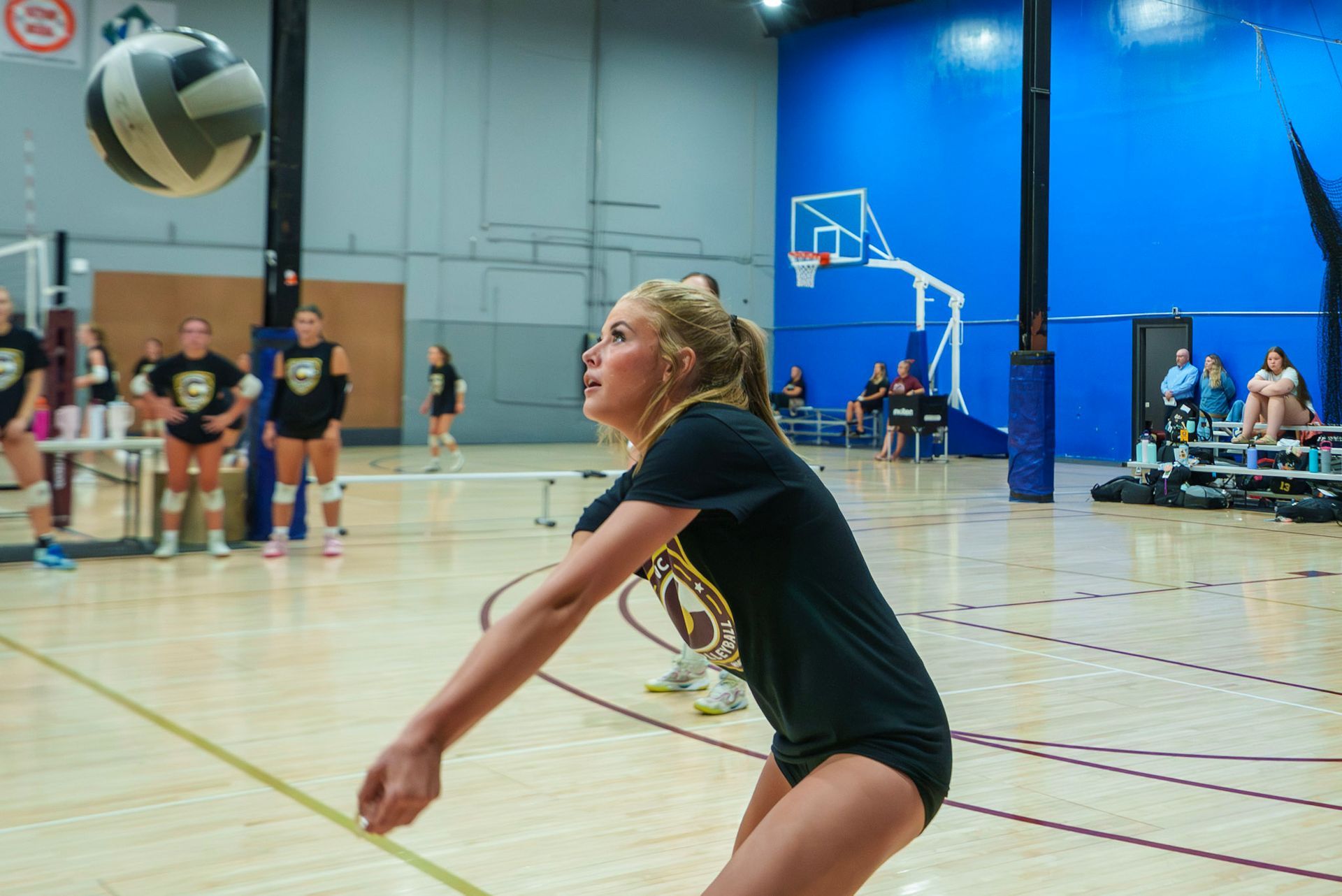 Female volleyball player in black uniform bumping the ball in a blue indoor court.