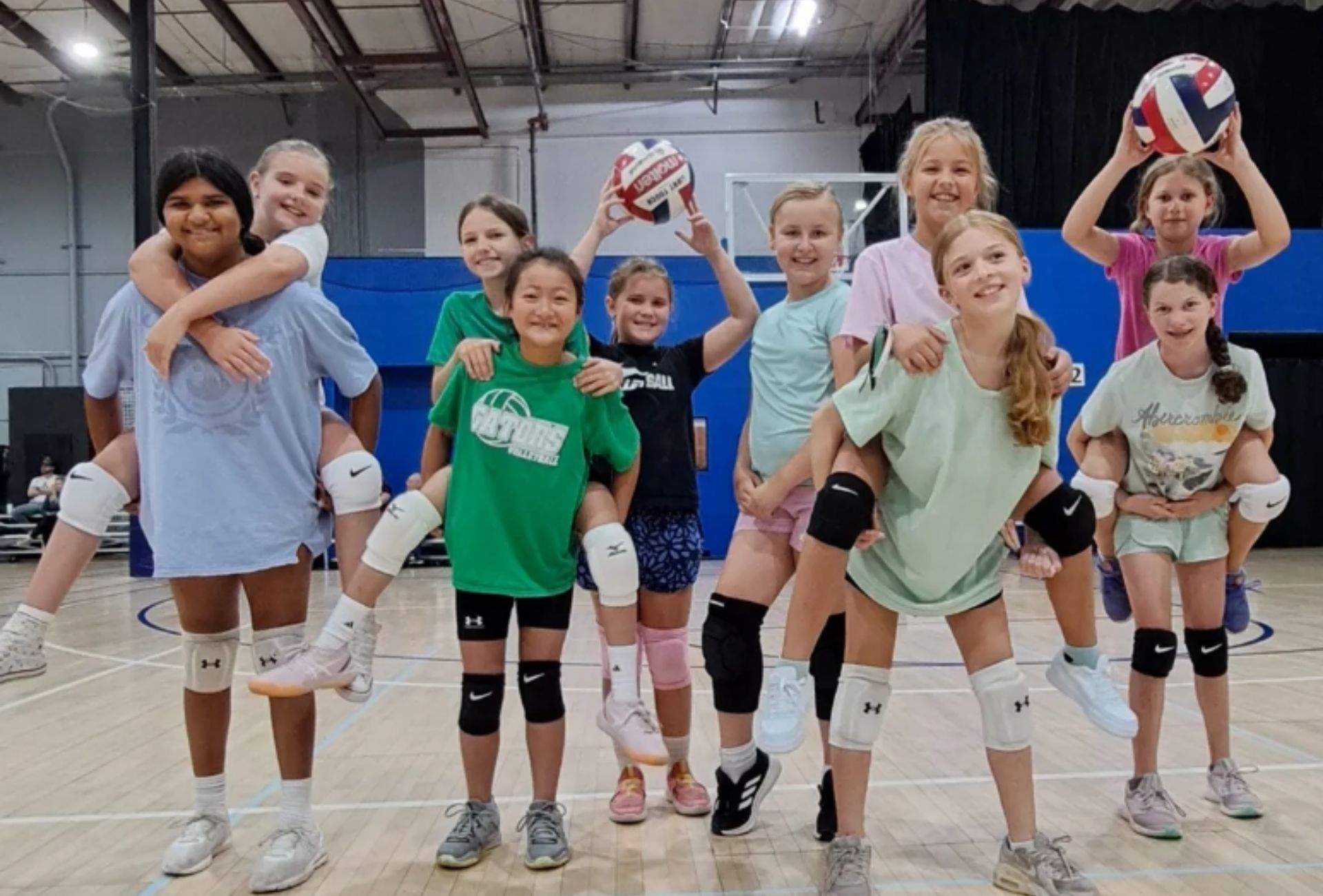 Girls, some carrying others piggyback style, pose with volleyballs in a gym, smiling.