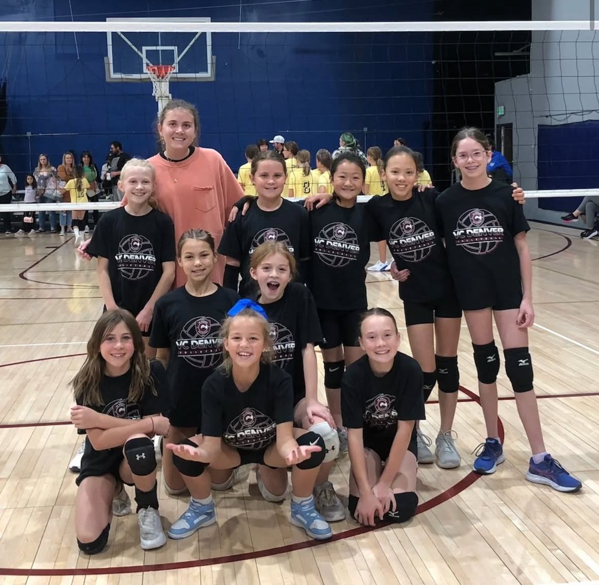 Youth volleyball team posing on a court, wearing black shirts, kneeling, and smiling.
