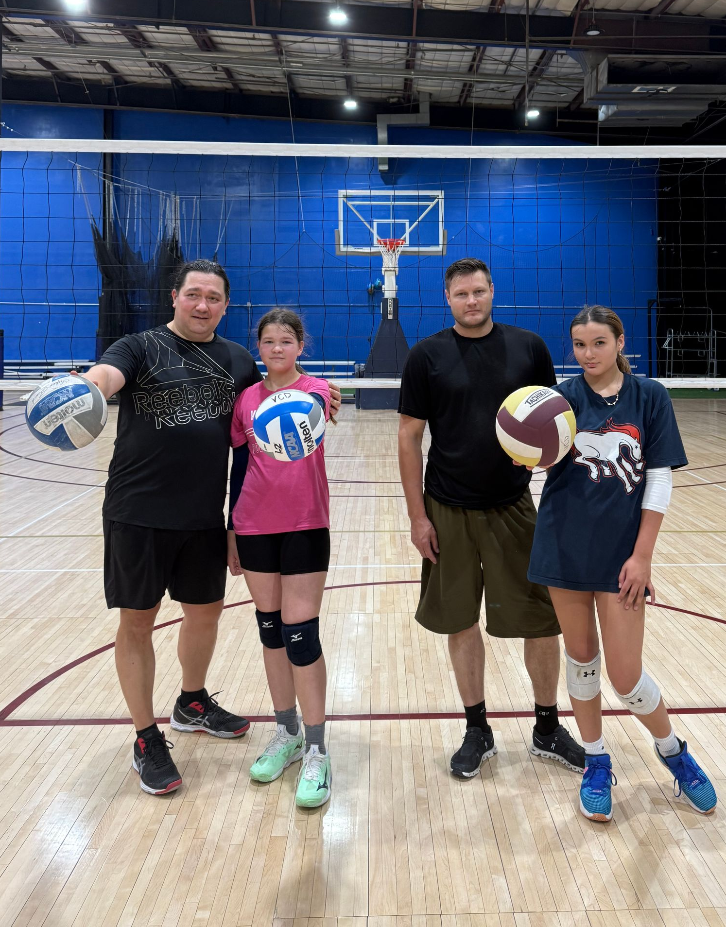 Four volleyball players posing in a gym, holding volleyballs in front of a net.