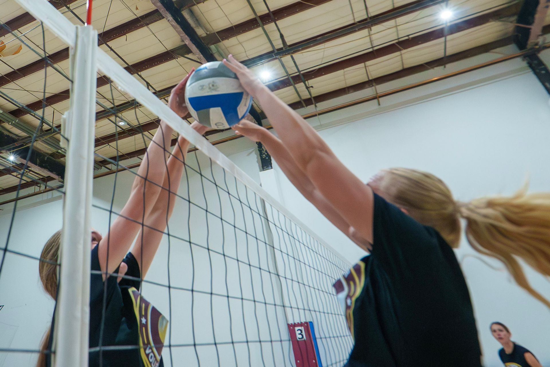 Two volleyball players blocking the ball near the net inside a gymnasium.