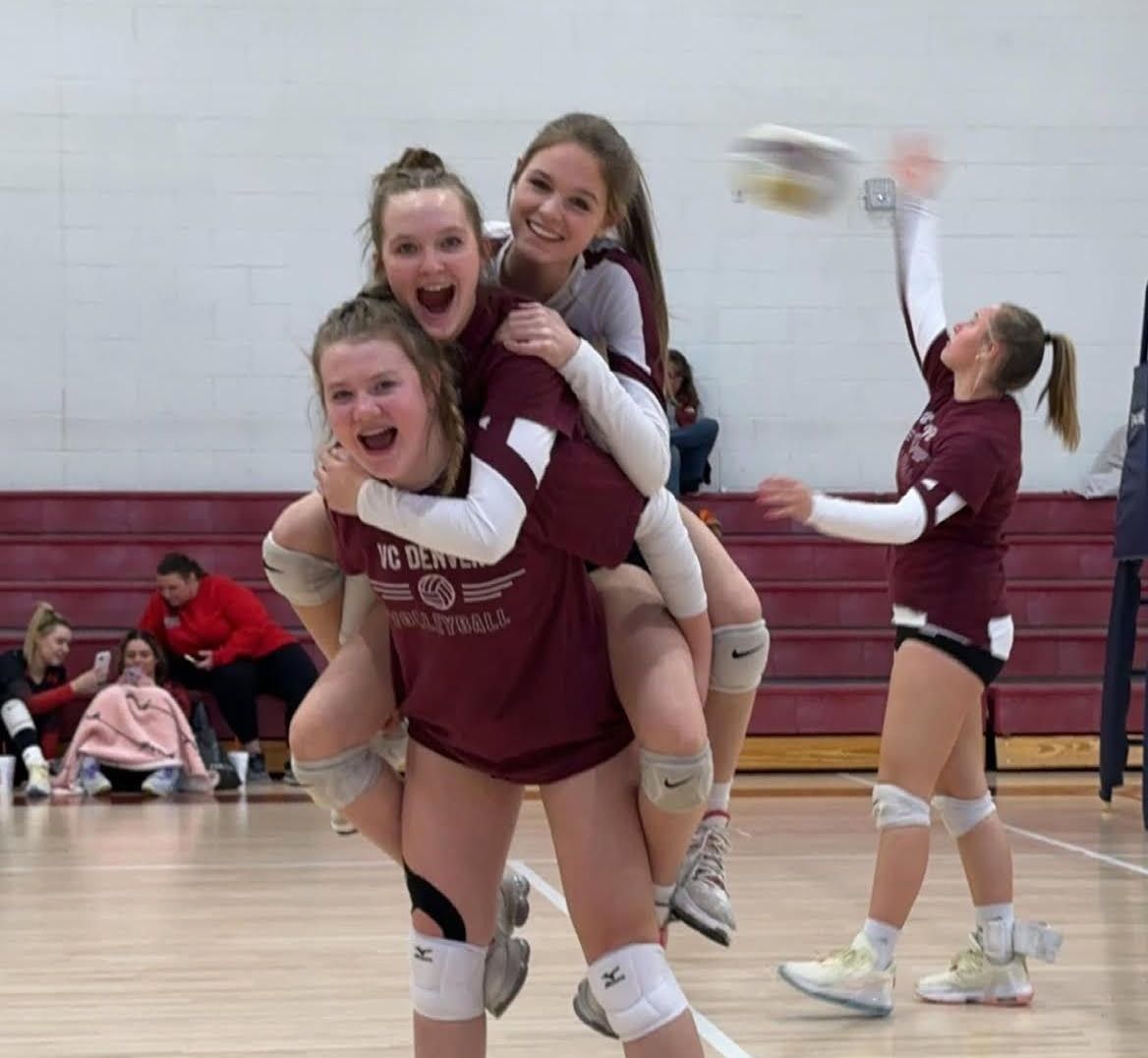 Volleyball players celebrate a point with piggyback rides. Two cheer while a third prepares to hit. Indoor court.