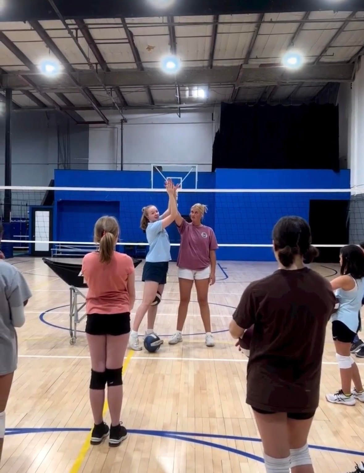 Two women high-five, volleyball court. Others watch. Indoors, blue walls, bright lights.