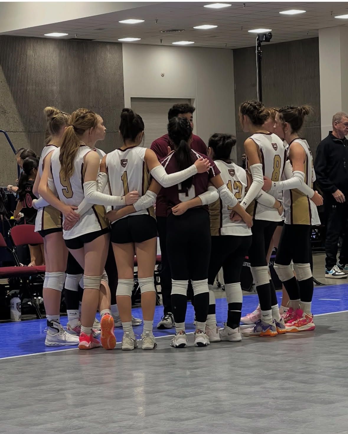 Volleyball team in a huddle, arms around each other, wearing black and white uniforms indoors.