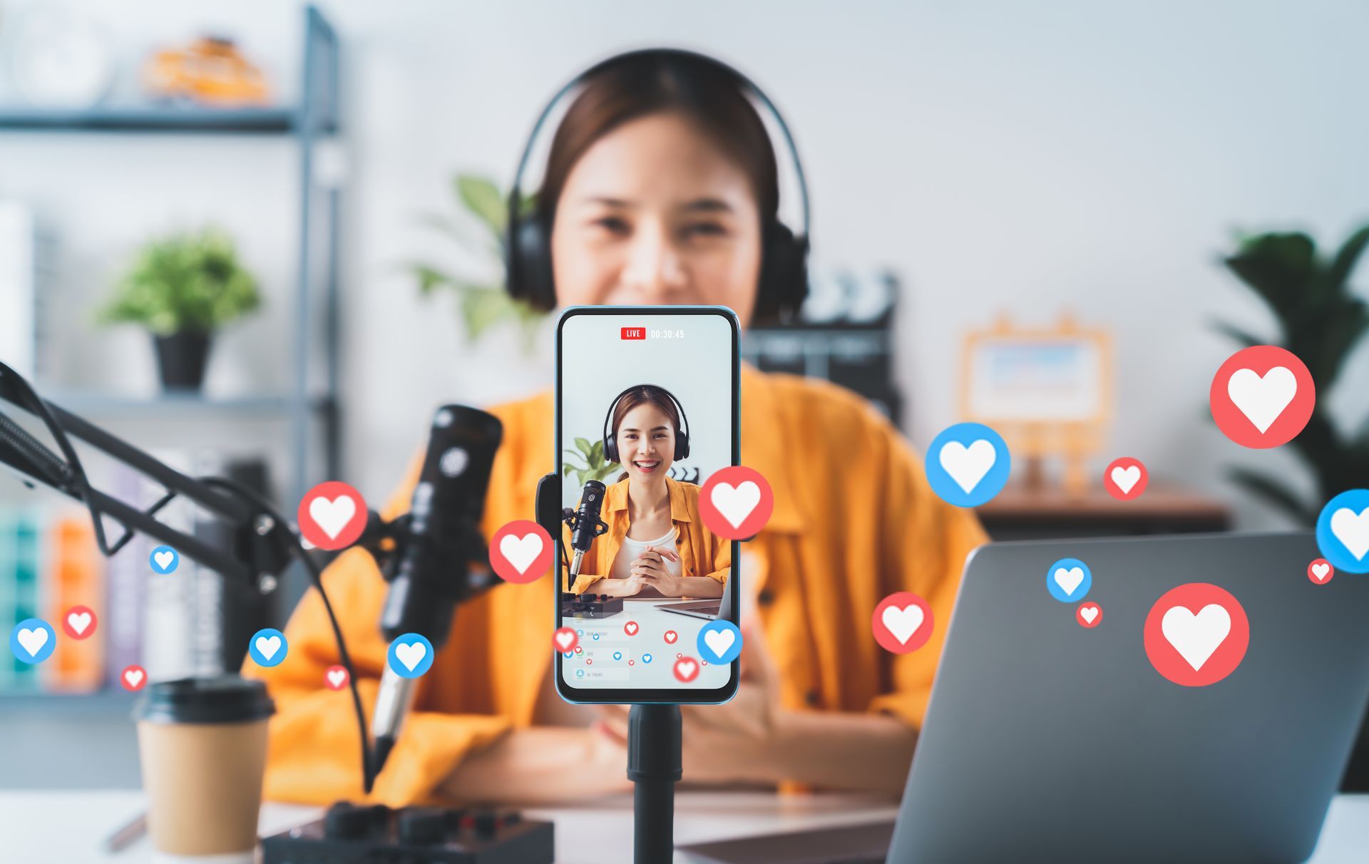 Woman live streams with phone and laptop, surrounded by microphone and coffee. Red and blue hearts float.