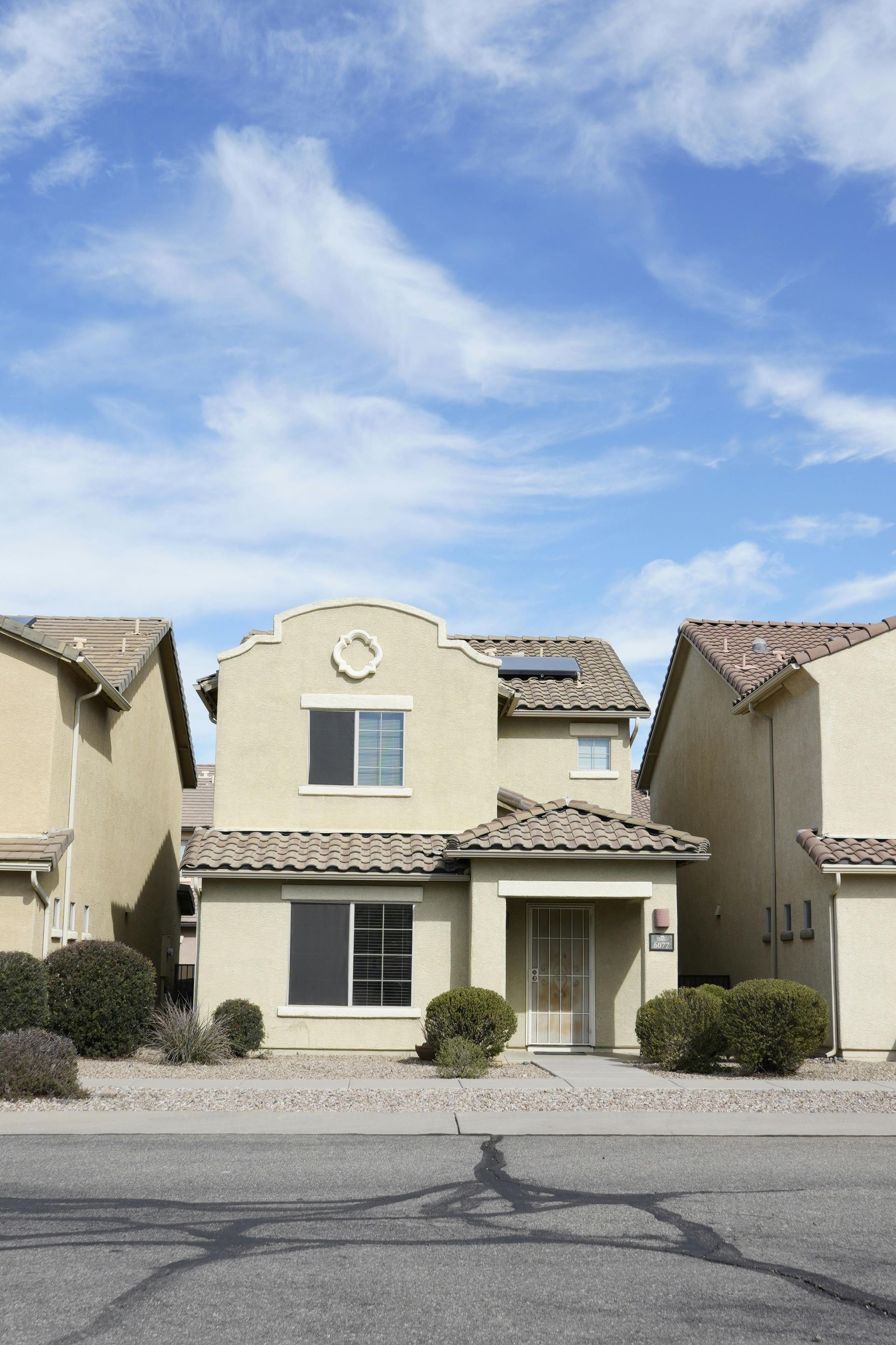 Two-story beige house with red tile roof, flanked by similar houses on a street, under a blue sky.
