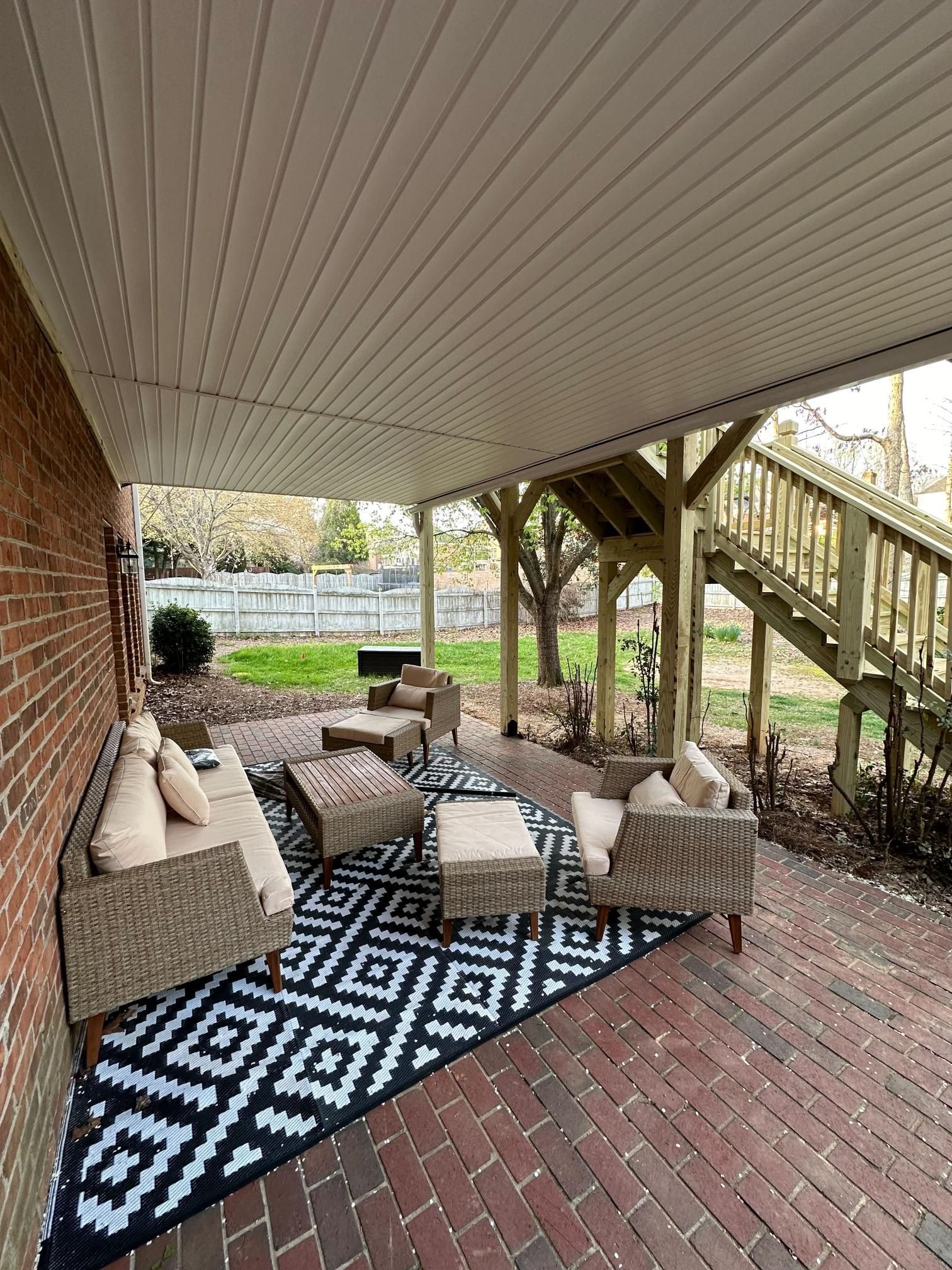 Covered patio with wicker furniture, black and white rug, red brick, and a wooden staircase.