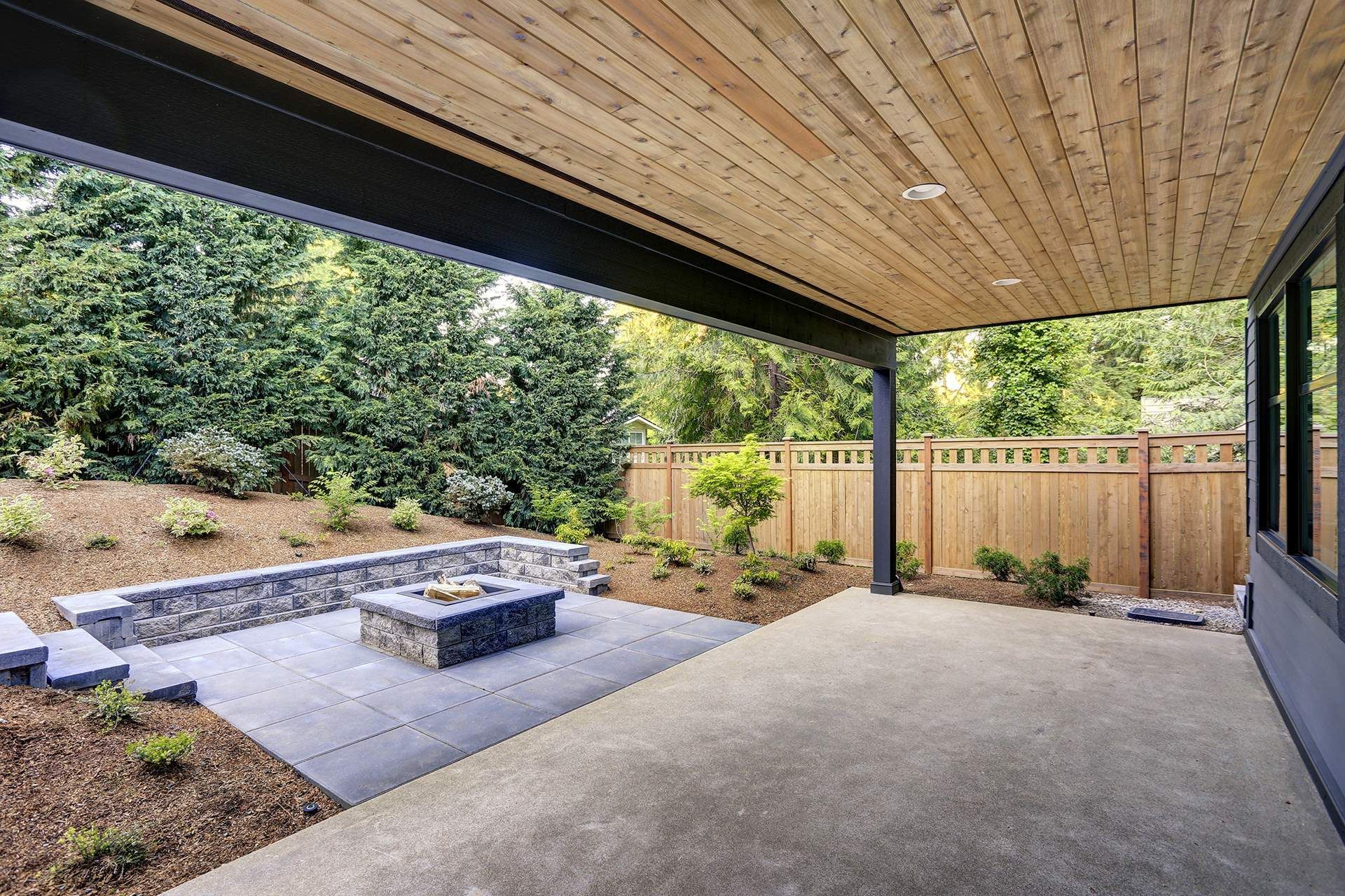 Covered patio with concrete floor, stone fire pit, and wooden ceiling, overlooking a landscaped yard.
