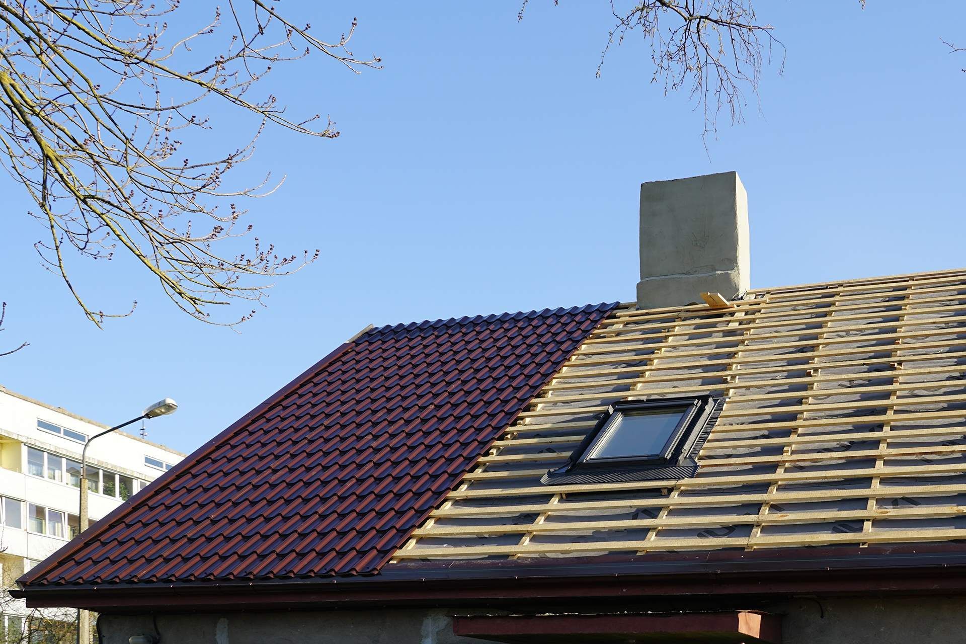 Partially finished roof with red metal tiles, wooden beams, and a skylight, near a chimney.
