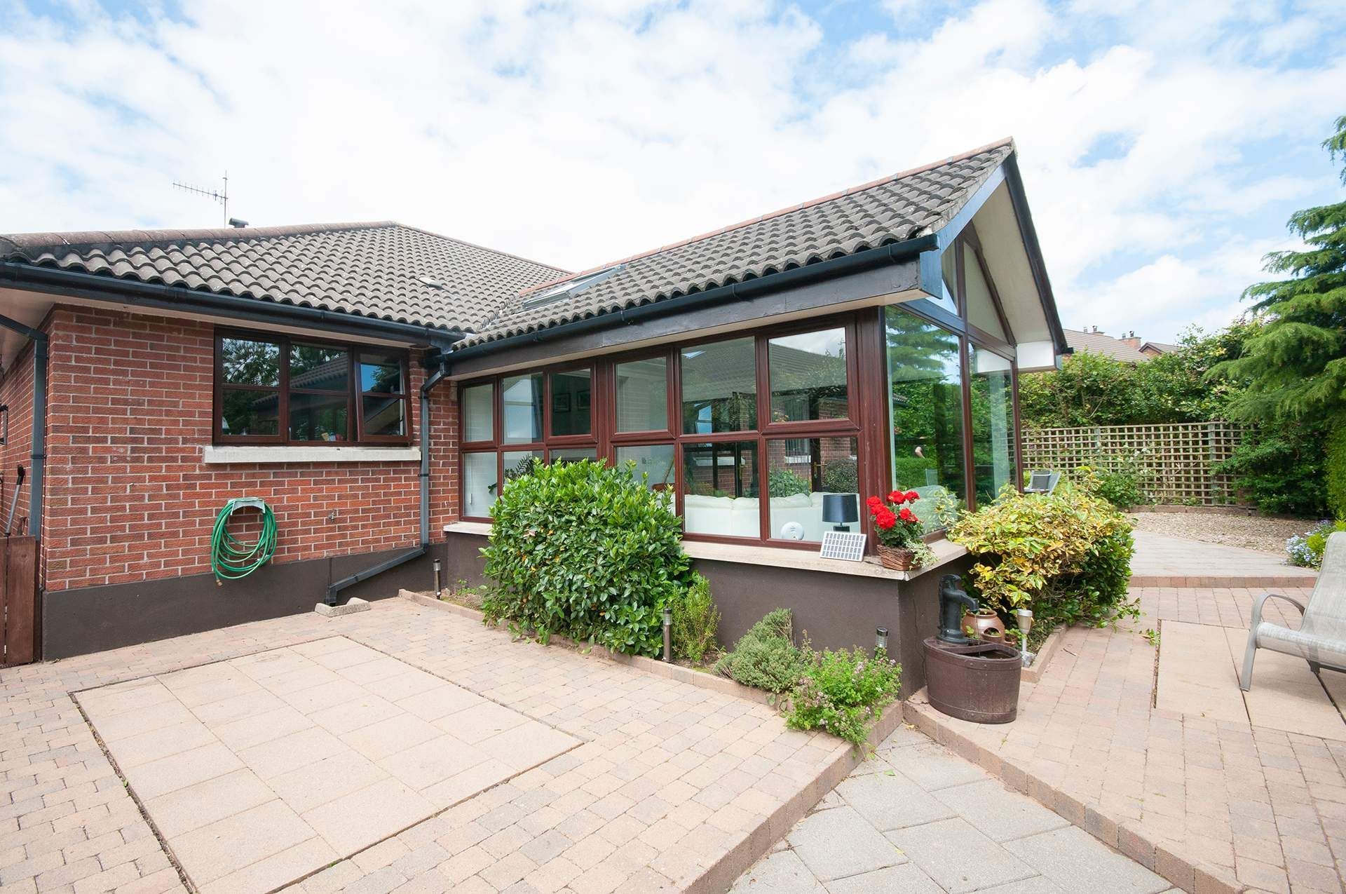 Brick house with a glass sunroom, brown trim, and a paved patio with greenery.