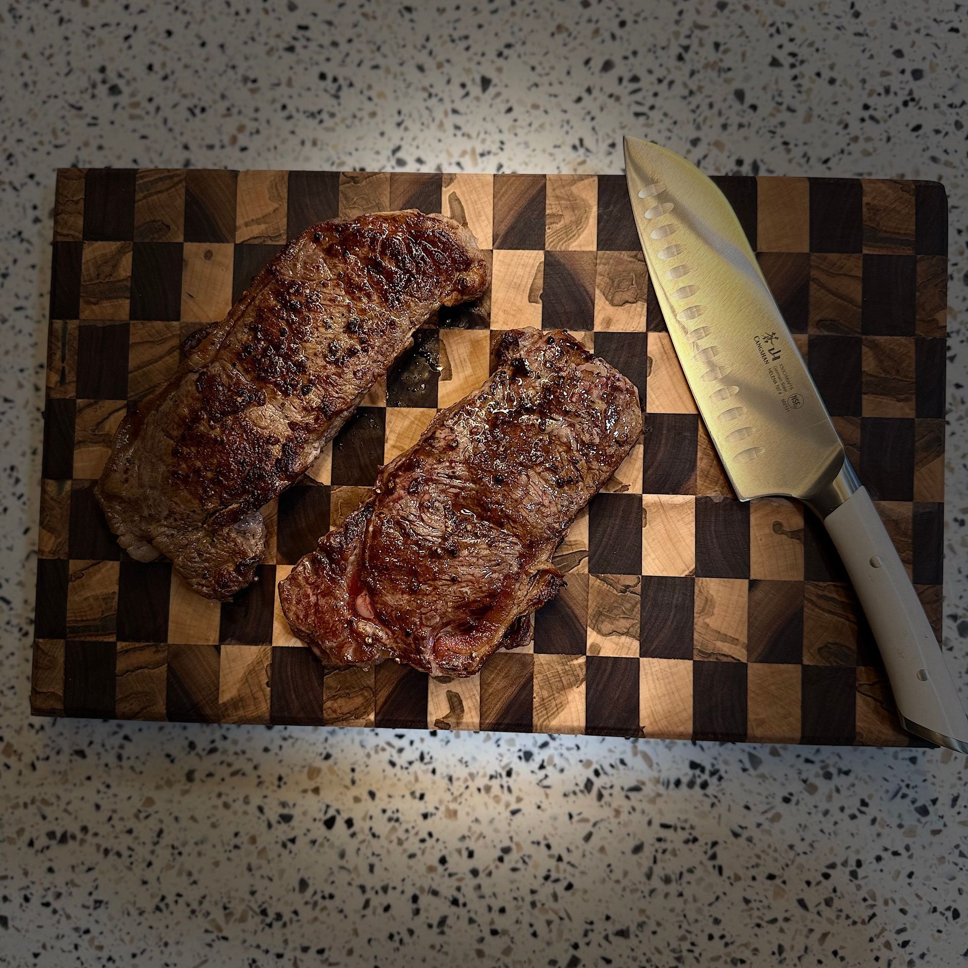 Two cooked steaks and a knife on a checkered cutting board.