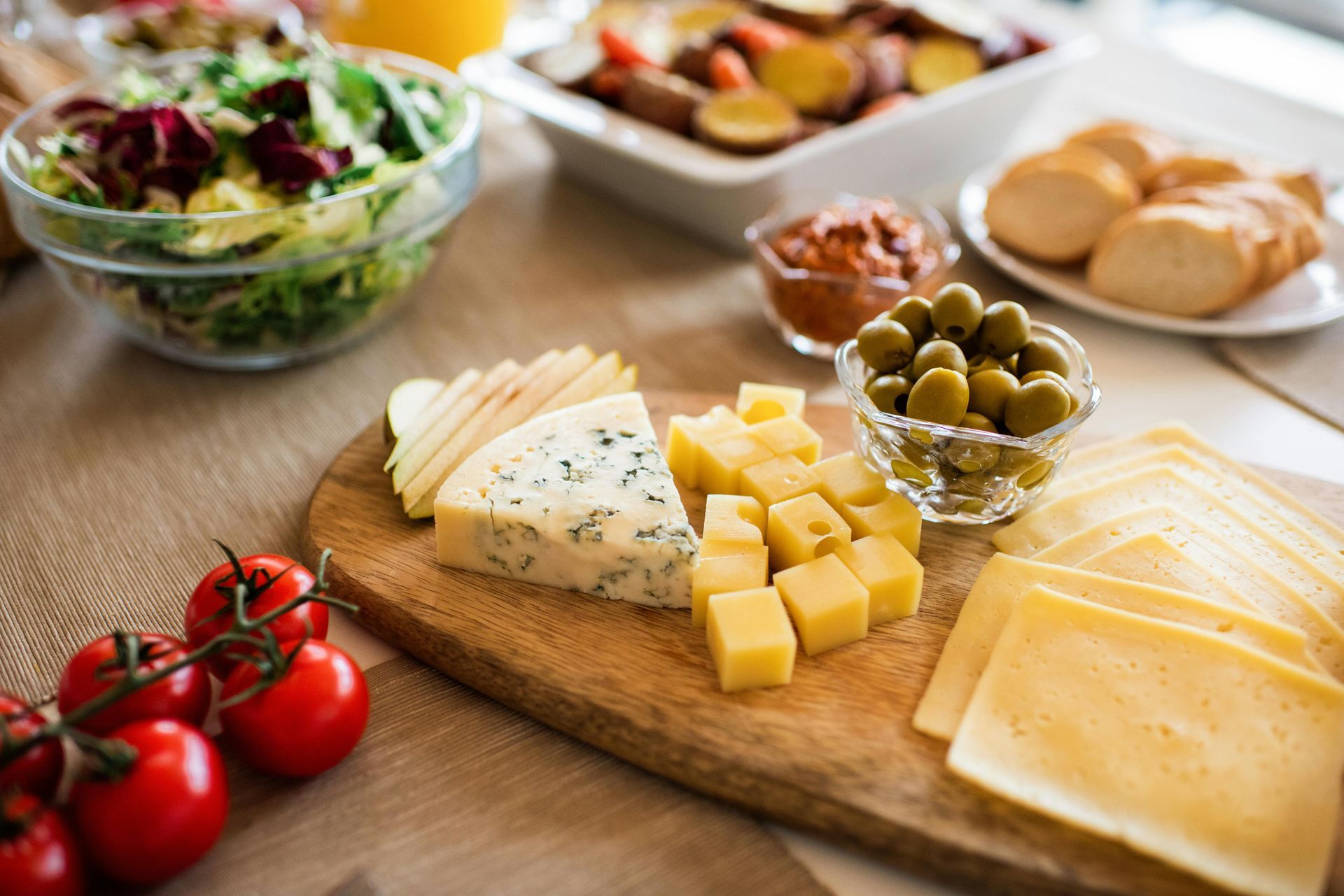 Wooden board with various cheeses, olives, salad, tomatoes, and bread on a table.