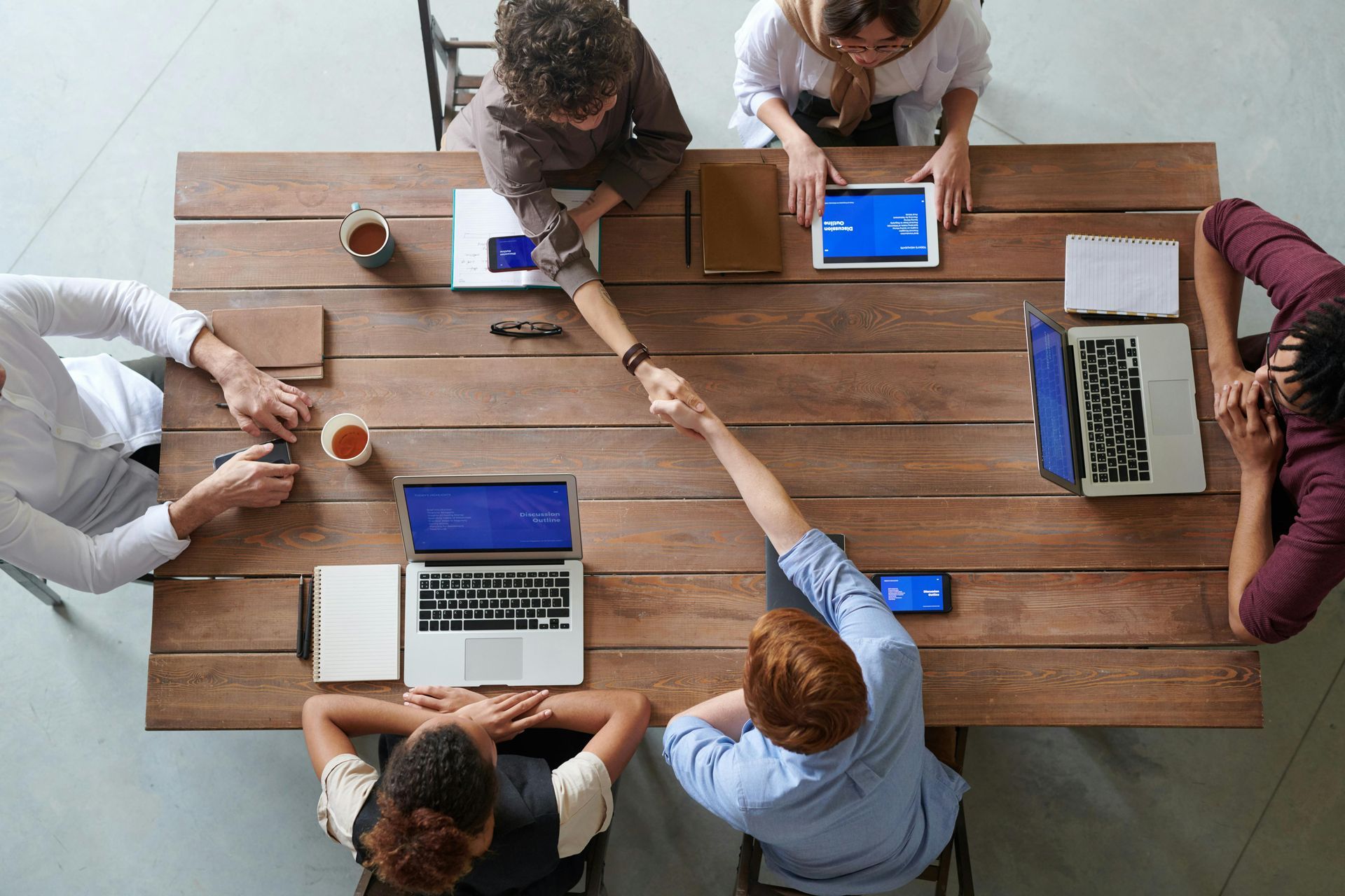 People shaking hands over a large wooden table with laptops, tablets, and coffee cups.