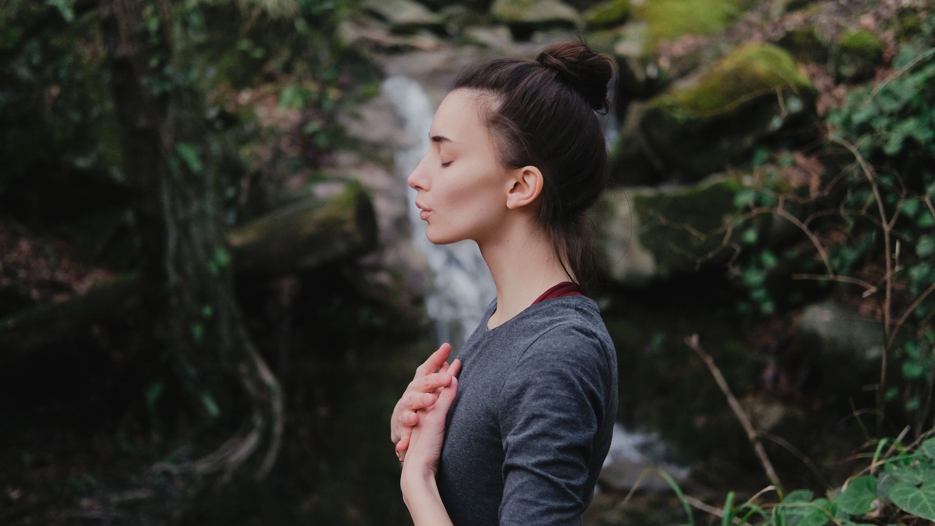 A woman is meditating in front of a waterfall in the woods.