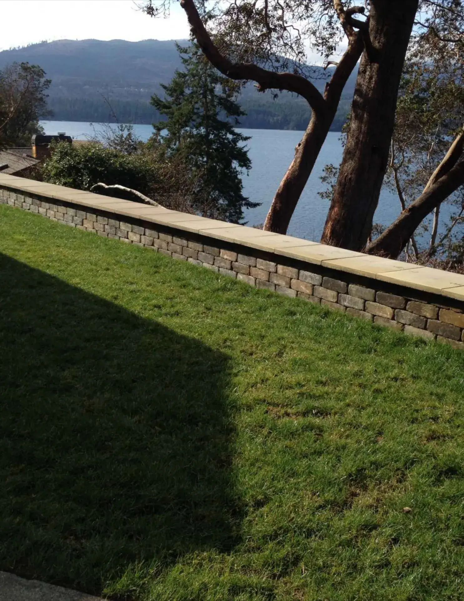 Grassy hillside beside a stone wall, overlooking a lake and distant mountains through trees.