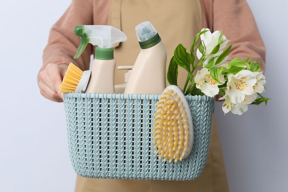 Person holding a blue basket filled with cleaning supplies, a brush, and white flowers. The person is wearing a tan apron.