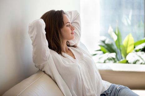 Woman relaxing on a couch, eyes closed, hands behind head. 