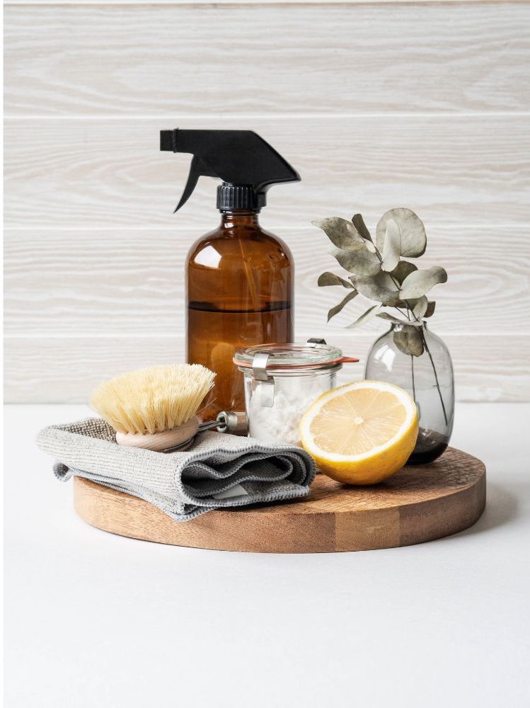Brown spray bottle, baking soda, lemon, brush, and cloth on a wooden tray, suggesting natural cleaning products.