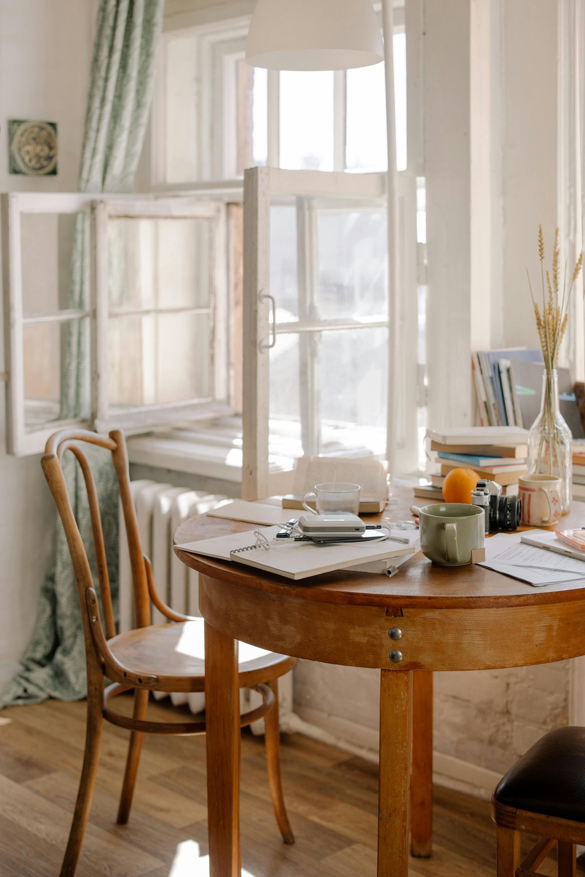 Wooden table set near a bright window; a chair is pulled up to the table cluttered with papers and a small orange.