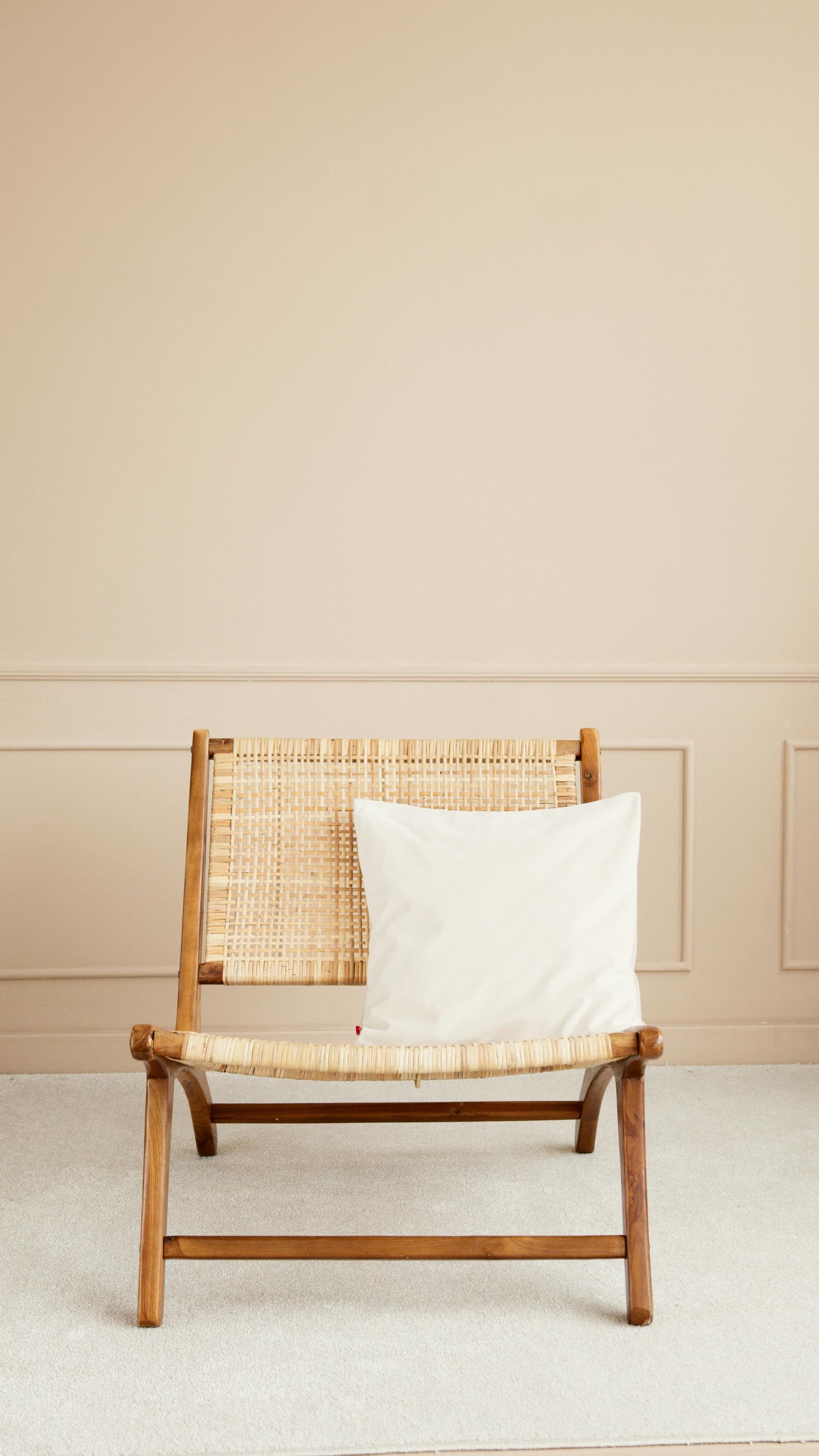 Wooden chair with woven back and seat, beige pillow, against a light-colored wall with molding; on a cream carpet.