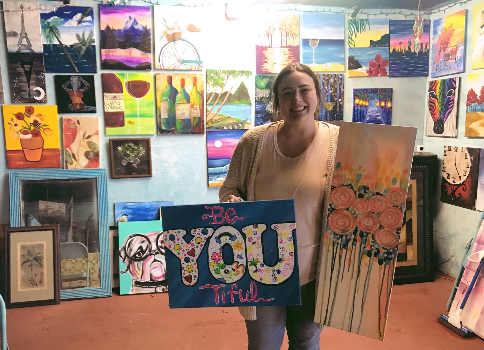 A woman is holding two paintings in front of a wall of paintings.