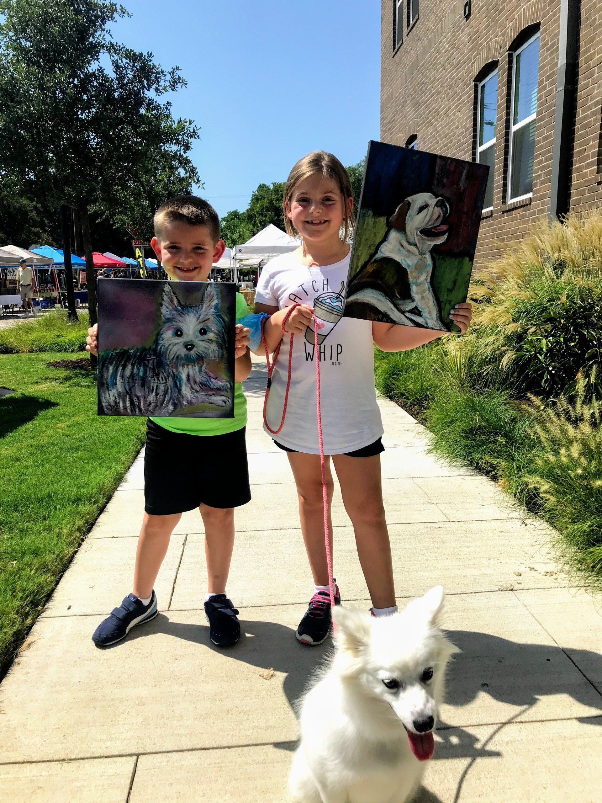 A boy and a girl are standing on a sidewalk holding paintings of their dogs.