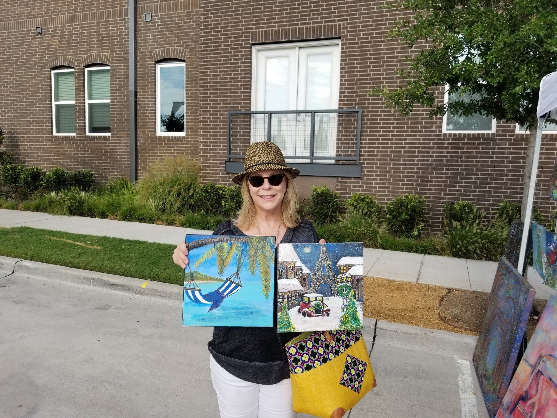 A woman is holding two paintings in front of a brick building.