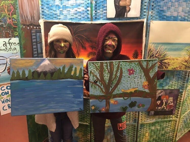 Two people are holding paintings in front of a wall.