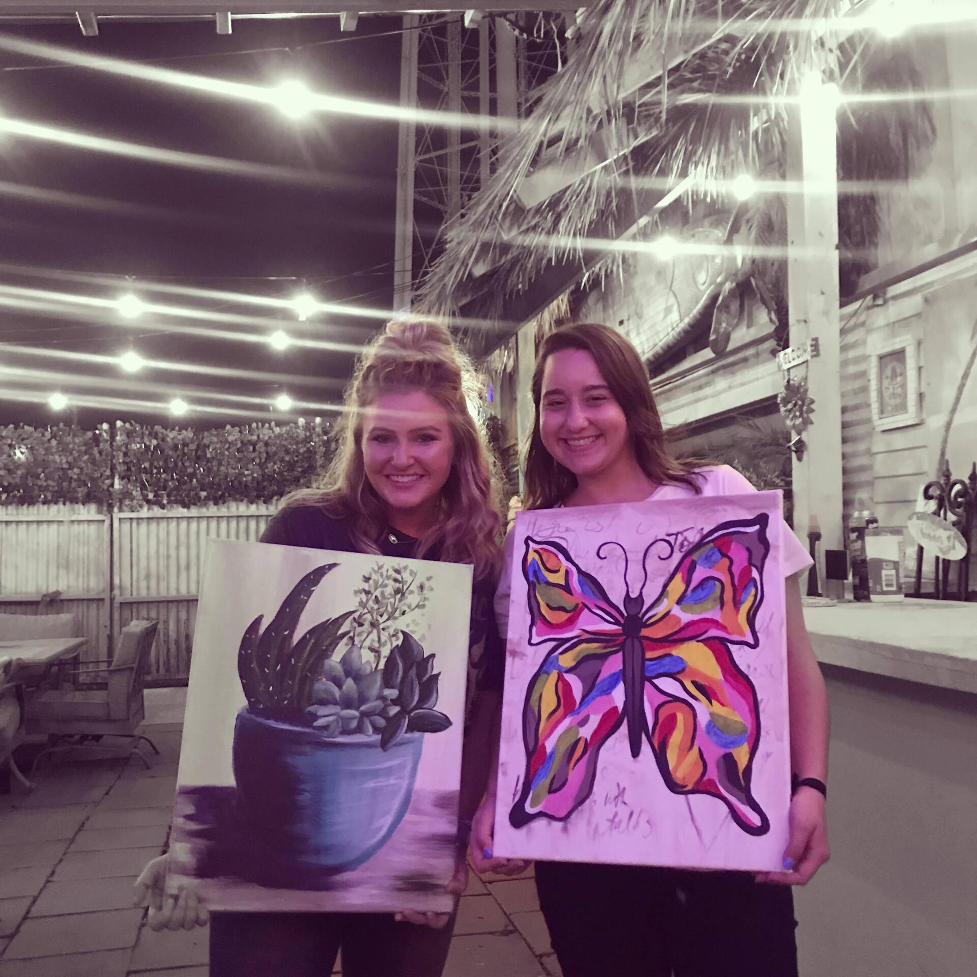 Two women are holding paintings of a butterfly and a potted plant